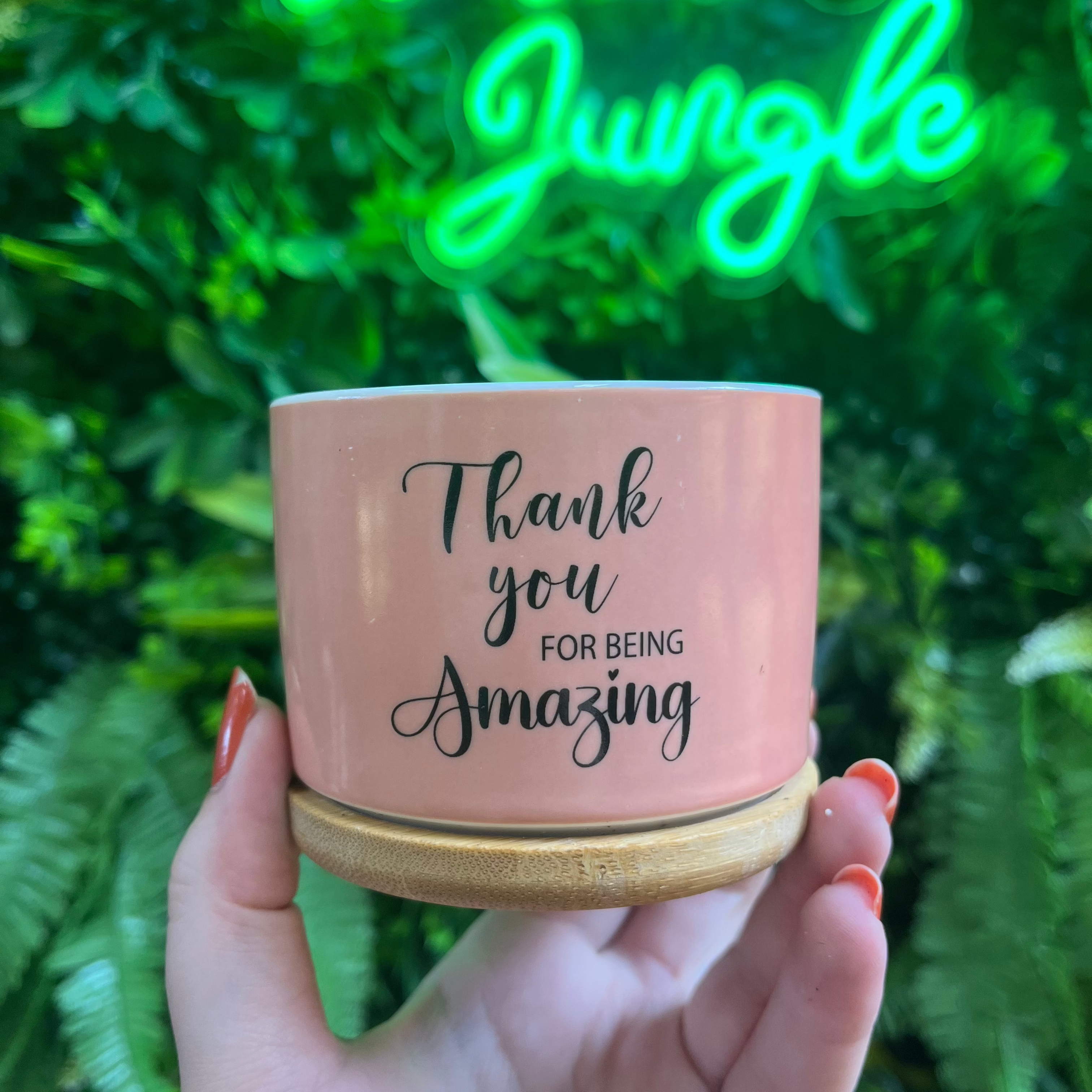 Pink mug with 'Thank you for being amazing' text held in front of a neon sign and green foliage.